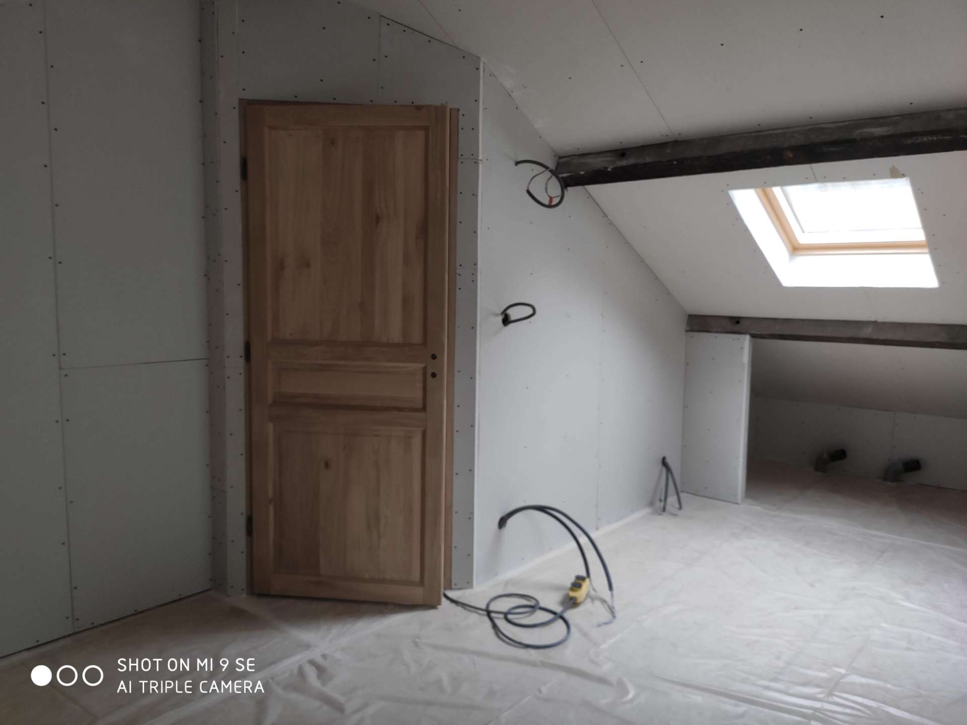 Combles en cours d'aménagement : porte en bois clair, murs de plaques de plâtre, poutre, fenêtre de toit et câbles électriques au sol.