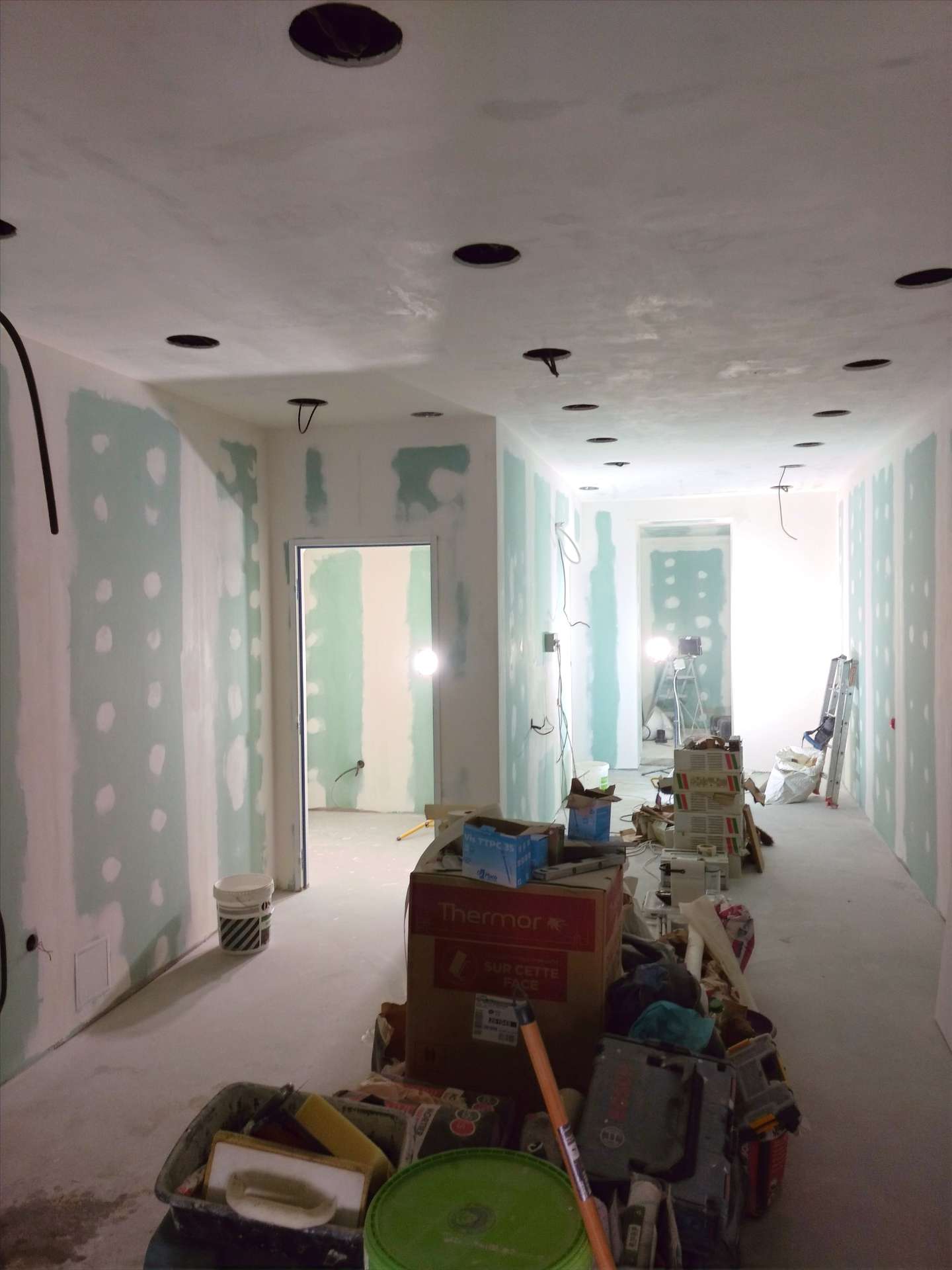 Couloir en travaux, murs et plafond percés de spots, cartons et outils empilés au sol.
