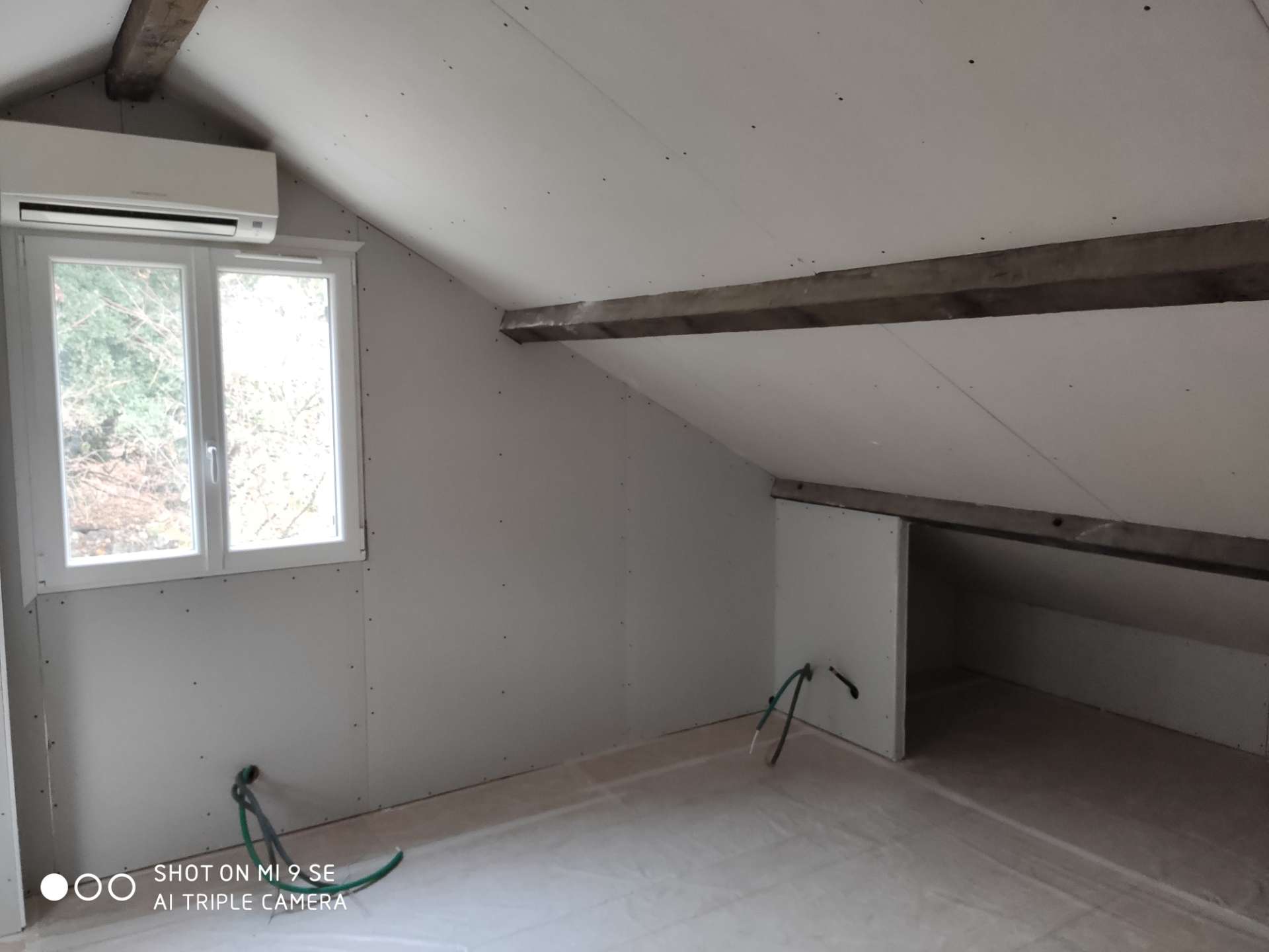 Combles en cours d'aménagement : plaques de plâtre au plafond et aux murs, fenêtre blanche à gauche, poutres apparentes et sol couvert d'une bâche.