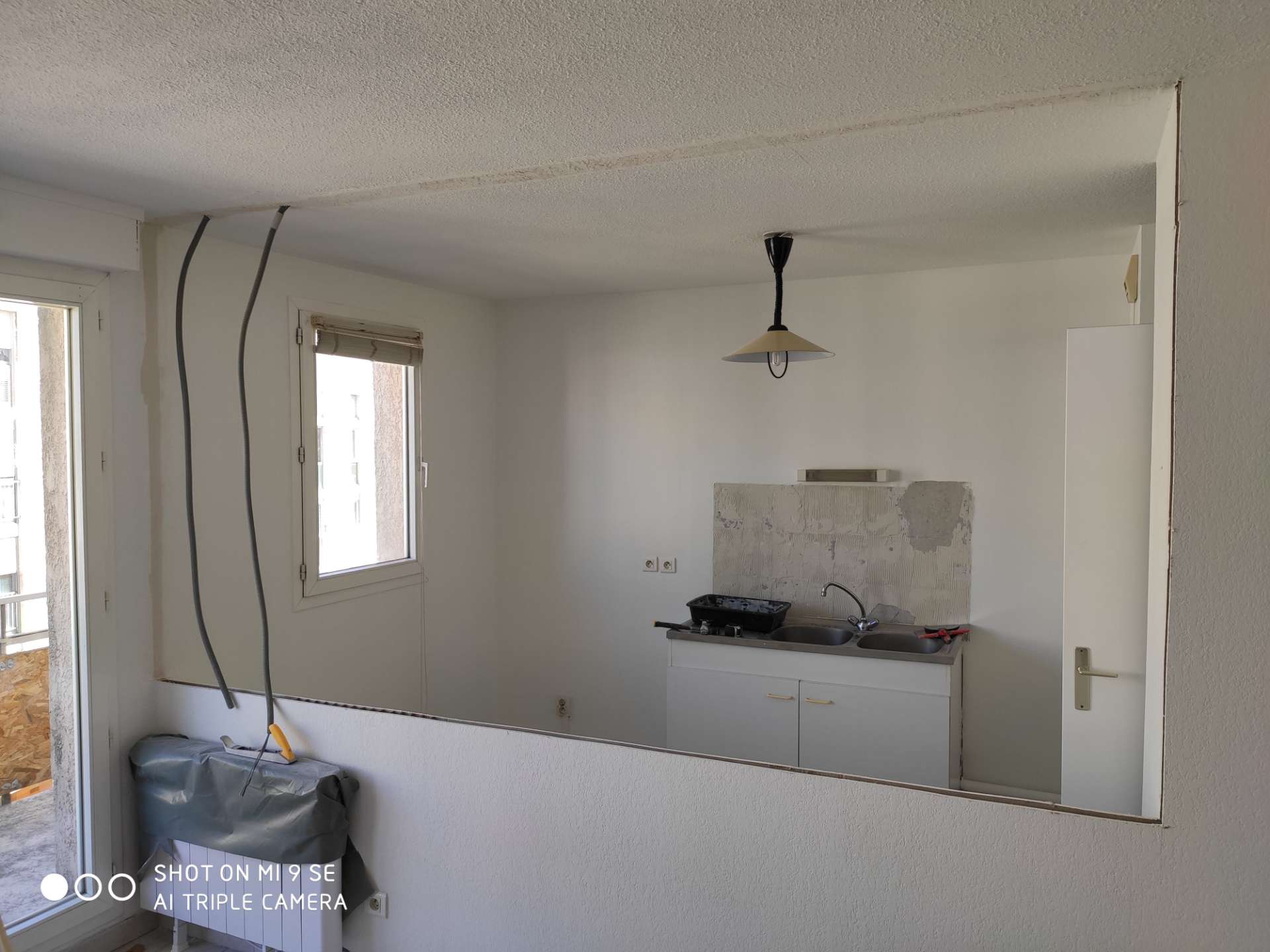 Pièce en travaux avec un grand miroir reflétant un évier double et une crédence, câbles pendant au mur et radiateur couvert d'une bâche.