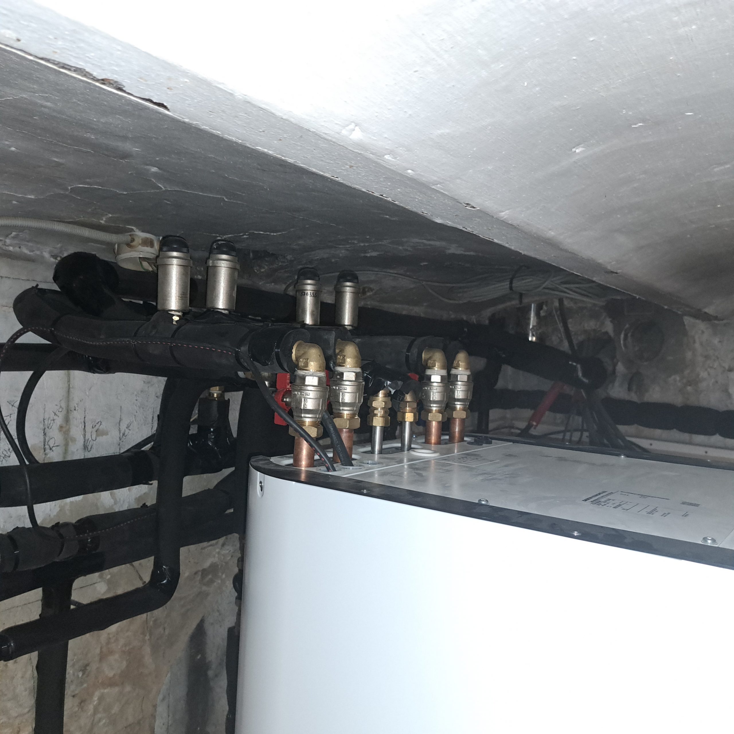 Installation de chauffage sous un plafond bas : tuyaux noirs isolés, vannes en laiton et cuivre au-dessus d'un caisson blanc.